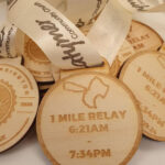 Wooden medals with ribbons feature a kangaroo design and engraved text reading 1 Mile Relay 6:21 AM - 7:34 PM 07.09.24. Medals are stacked, some showing partial logos and text.