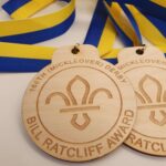 Wooden medals with a fleur-de-lis design, labeled "166th (Mickleover) Derby Bill Ratcliff Award," attached to blue and yellow ribbons.