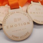 Wooden medals with orange ribbons and engraved text, including "EMOTION FITNESS HUB" and various names.