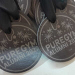 Close-up of multiple round medals with a black ribbon, engraved with palm tree designs and the text PUREGYM ALUMNI 2023. The medals are placed on a flat surface.