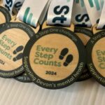 Close-up of several wooden medals with the text Every Step Counts 2024 and Supporting Devons mental health on a white surface. They have white and green lanyards attached.