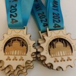 Wooden medallions with a gear shape and a design of the Brighton Pavilion, attached to blue ribbons with the text LONDON TO BRIGHTON CYCLE RIDE 5TH MAY 2024.