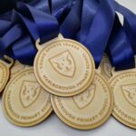 A bunch of medals with blue ribbons are displayed. The wooden medals are engraved with Young Sports Leader and Marlborough Primary School, along with a shield and banner emblem.