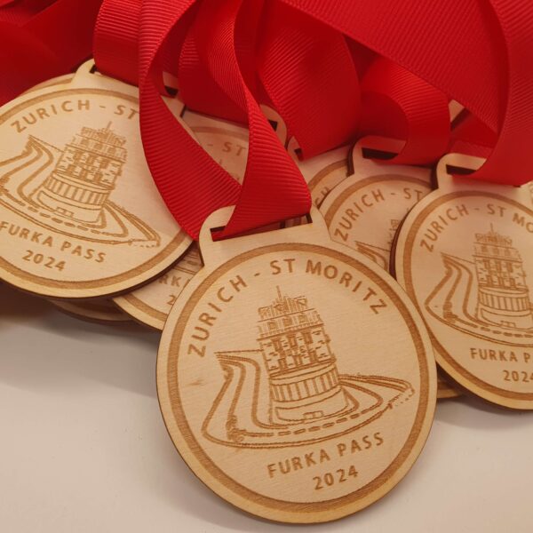 Wooden medals with red ribbons are stacked together. Each medal is engraved with Zurich - St Moritz and Furka Pass 2024, along with a design of a winding road and tower.