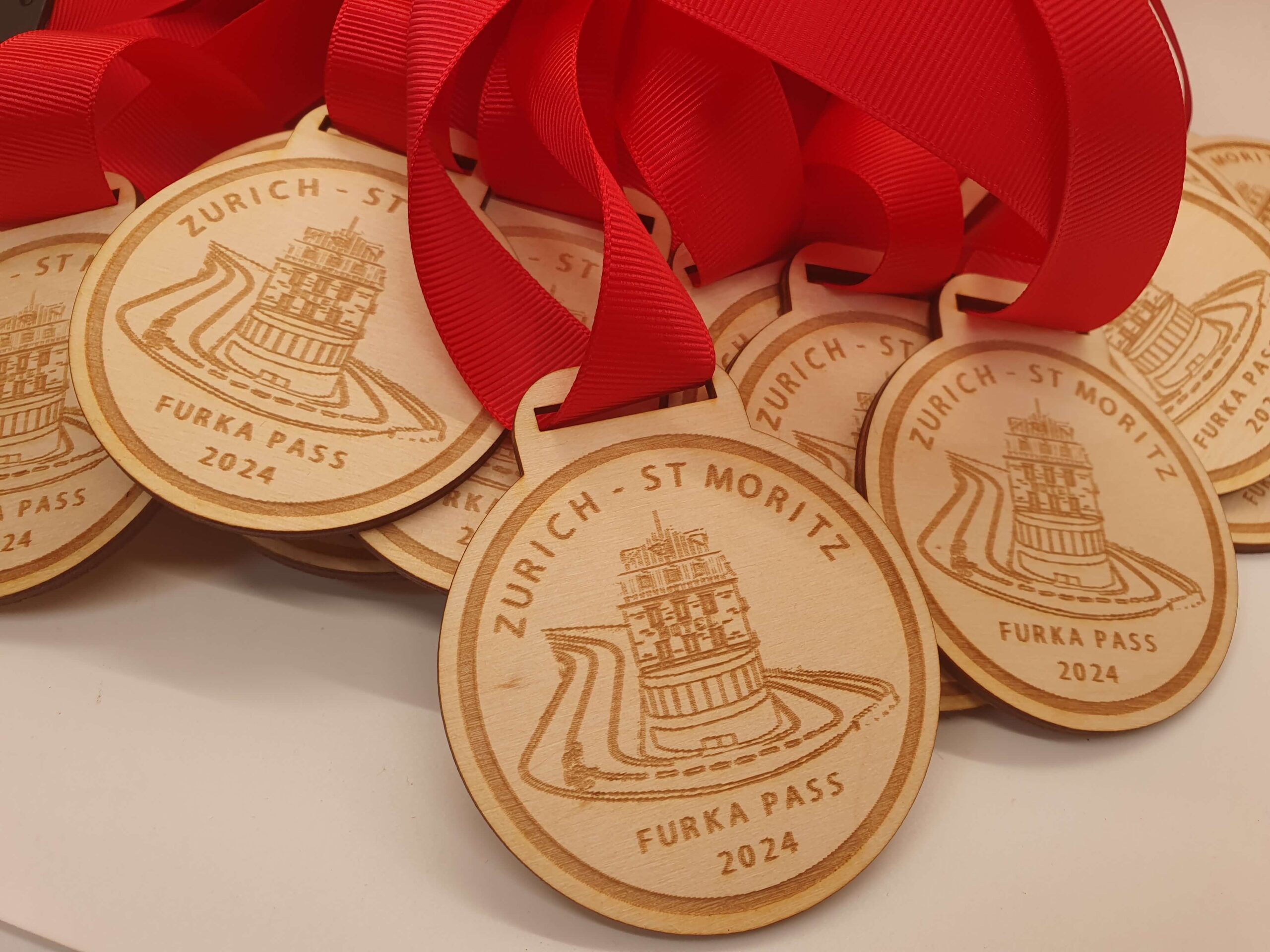 Wooden medals with red ribbons are stacked together. Each medal is engraved with Zurich - St Moritz and Furka Pass 2024, along with a design of a winding road and tower.