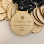 A pile of wooden medals with black ribbons, each engraved with THAKEHAM, a lion emblem, and the text Horsham Rugby Club Minis 2024.