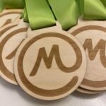 Wooden medals with a carved M inside a circle, attached to light green ribbons, arranged in a row on a white surface.