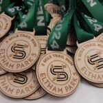 A pile of wooden medals with 5 Mile Trail Run Eltham Park 5 inscribed on them, displayed with green ribbons marked 2024.