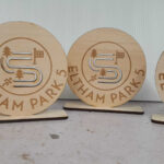 Four circular wooden plaques on stands, engraved with the text ELTHAM PARK 5 and a design featuring a stylized path, trees, and a flag.