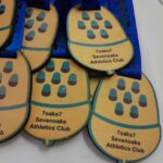 Medals with a wooden finish and blue ribbons are displayed. The medals feature a footprint design and are labeled 7oaks7 Sevenoaks Athletics Club. A piece of paper is partially visible in the background.