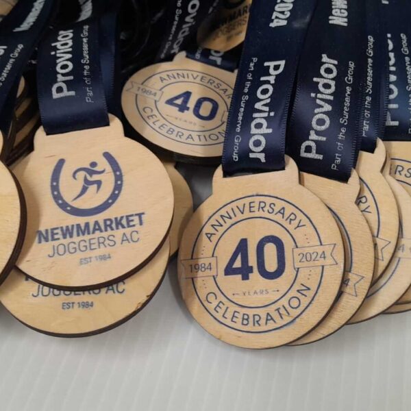 A collection of wooden medals with blue ribbons, featuring the logo of Newmarket Joggers AC. The medals commemorate the 40th anniversary celebration of the club, with text indicating ANNIVERSARY CELEBRATION and dates 1984-2024.