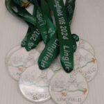 A collection of green lanyard medals labeled Lingfield 10s 2024 with illustrations of runners. The medals are designated for different categories like 1st Male and 1st Female 45+ placed on a flat surface.