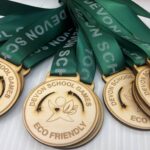 Wooden medals with a green ribbon are labeled Devon School Games and Eco Friendly, featuring an engraved bird logo. The ribbons are laid out, displaying the embossed text Devon School multiple times.