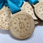 A close-up of wooden medals with blue ribbons, engraved with Bible Society 220th Anniversary 2024 Thank You and a logo in the center, resting on a white surface.