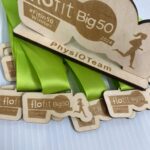 Wooden medals with green ribbons featuring flofit Big50 2024 branding. A larger medal displays #Fitin50 Winner and a silhouette of a running figure, with PhysiOTeam inscribed. Medals are laid out on a white surface.