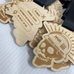 A pile of wooden medals featuring a skull design with a helmet. The text on the medals includes event details and locations, with phrases like Summer Maker Gear” and “Print It Forward.” Each medal has a black ribbon attached.