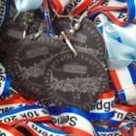 A close-up of several medals with the word Finisher and a mountain design, attached to ribbons. The ribbons are red, white, and blue with text Severn Bridges 10K 2024. A light blue ribbon is also visible.