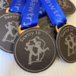 Close-up of round black medals with blue ribbons labeled Sawtry 10. The medals feature the silhouette of two running figures.