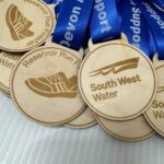 A pile of wooden medals with blue ribbons. The medals are engraved with a running shoe and the text Reservoir Run Fun and South West Water.