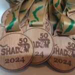 Wooden medals with 50 Years of Shadow 2024 engraved on them are displayed. They have gold ribbons with a green pattern.