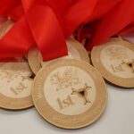 A close-up of multiple 1st place medals from the Welsh Field Archery Association Indoor Championship. The medals are wooden with a design featuring a dragon and a bullseye target, attached to red ribbons.