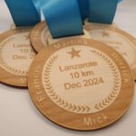 Three wooden medals with blue ribbons are engraved with laurel designs, a star, and the text Lanzarote 10 km Dec 2024. Names Martina, Derrick, Francis, and Mick are visible around the edges.