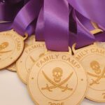 A close-up of several wooden medals engraved with FAMILY CAMP 2025 and a skull with crossed swords, each hanging from a purple ribbon.