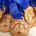 A close-up of several wooden medals with blue ribbons. The medals are engraved with a compass design, a figure holding a map, and the text SKAISTIS ORIENTEERING CLUB.