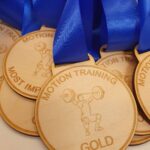 A close-up of wooden medals with blue ribbons. The medals are engraved with a weightlifter image and texts like “MOTION TRAINING,” “GOLD,” and “MOST IMPROVED.”.