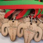 A close-up of several wooden bicycle-themed medals shaped like an elephant head, each with a red and green ribbon. The medals have YOUNGS and FINISHER engraved on them.