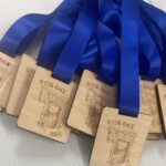 Several wooden medals with blue ribbons, engraved with NESK RACE 2025 and an illustrated design, are displayed on a white surface.