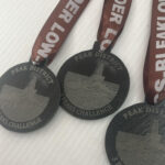 Three dark medals with brown ribbons, each engraved with PEAK DISTRICT 3 PEAKS CHALLENGE and an illustration of a hill or rock formation. The ribbons read KINDER LOW and BLEAK LOW. Medals are placed on a white surface.