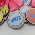 Several wooden medals with pink ribbons are shown. The medals are labeled School Games with gold, silver, and bronze centers, and text indicating GOLD, SILVER, and BRONZE MEDAL WINNER.