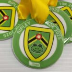Four green medals with yellow ribbons featuring a shield design, a smiling frog face, and a flower. The text RIPLEY 10K 2025 and the Liniar logo are visible on the medals.
