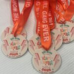 A group of round medals with orange ribbons reading THE WORST GIRL GANG EVER. The medals display 10 km, girl gang ever foundation 2025, and WE DO HARD THINGS in colorful text with a lightning bolt design.
