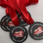 Four black medals with red ribbons, labeled Fidra Lions Hockey Club East Div 1 Champions 2015, featuring a white lion silhouette over a red splash graphic on a textured white surface.