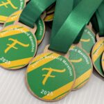 A close-up of wooden medals with green and yellow designs, green ribbons, and the text Fircroft Backyard Marathon 2025 printed on each medal. The medals are stacked on a white surface.