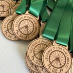 A pile of wooden medals with green ribbons. The medals are engraved with a bicycle wheel, runner icons, and text that reads Legging It for the Library 5K Fun Run.