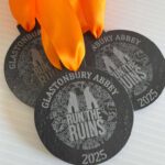 Three round black medals with orange ribbons. The medals read GLASTONBURY ABBEY RUN THE RUINS 2025 with an illustration of ruins and decorative patterns in the background.