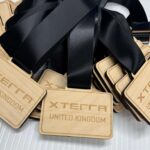 A pile of rectangular wooden medals with “XTERRA UNITED KINGDOM” engraved on them, each attached to a wide black ribbon. The medals are stacked on a white surface.