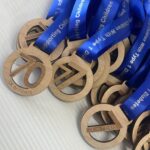 A pile of wooden medals with blue ribbons printed with text. The medals are engraved with 7K IN 7 DAYS and have circular cutouts. The medals rest on a white textured surface.
