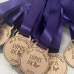 A group of wooden medals with purple ribbons, engraved with “26.2 Challenge LUPUS UK 2025” and a butterfly symbol, arranged on a white surface.