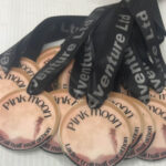 A pile of round medals with Pink moon and Ladies trail half marathon written on them, featuring a pink moon design and attached to black lanyards printed with Venture Ltd in silver letters.