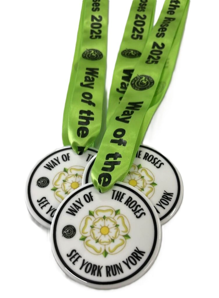 Three white Eco Medals with a yellow and green rose design and the words WAY OF THE ROSES SEE YORK RUN YORK are attached to bright green ribbons that read Way of the Roses 2025 in black text.