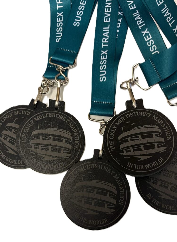 Five dark Eco Medals with teal ribbons reading SUSSEX TRAIL EVENTS. The medals are engraved with a building and the text THE ONLY MULTISTOREY MARATHON IN THE WORLD! on a white background.