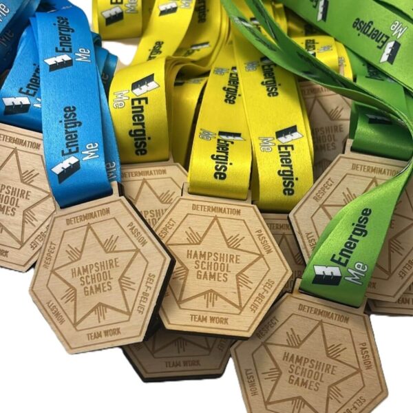 A pile of wooden hexagonal medals with blue, yellow, and green ribbons labelled Energise Me. The medals are engraved with Hampshire School Games, DETERMINATION, and TEAM WORK.