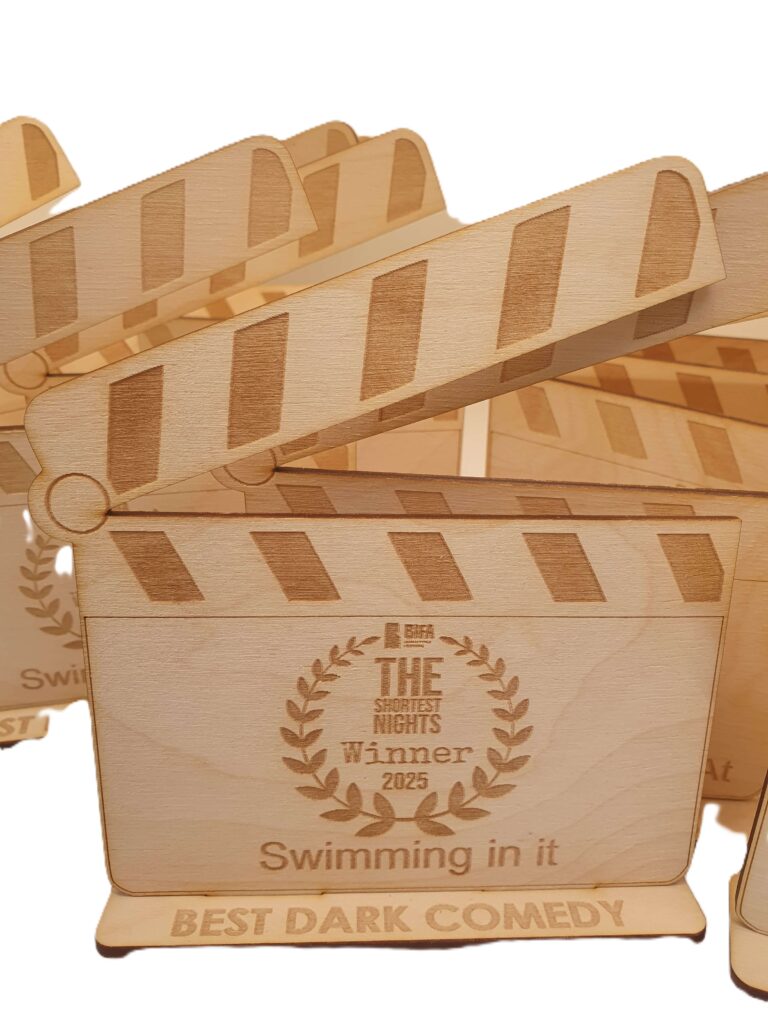 A wooden film clapperboard trophy, part of the Eco Medals collection, is engraved with “THE SHORTEST NIGHTS Winner 2025,” “Swimming in it,” and “BEST DARK COMEDY.” Several similar trophies are visible in the background.