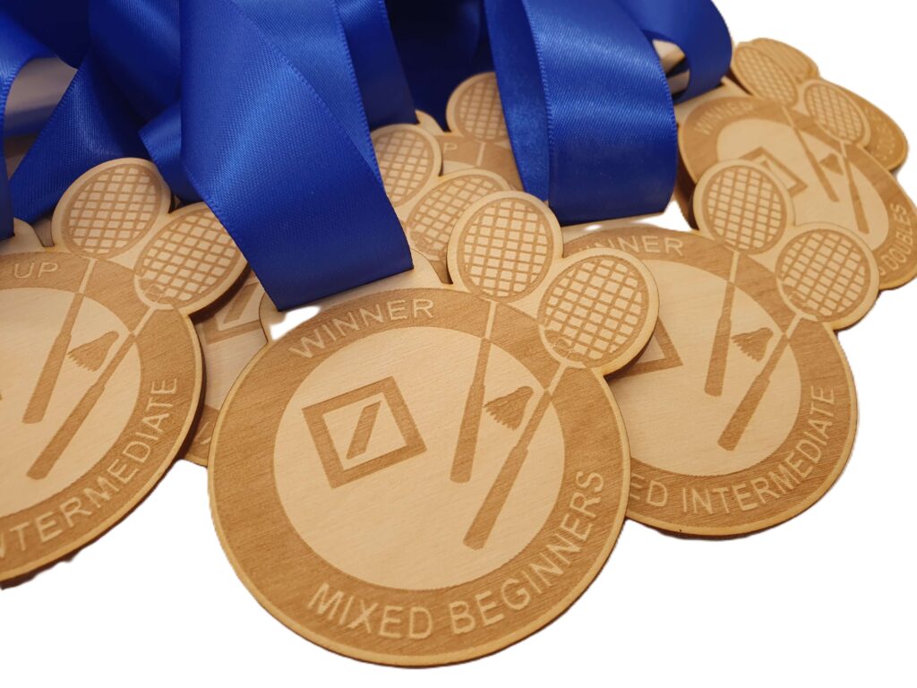 Close-up of several sustainable wooden medals made in the UK, each with blue ribbons. The medals feature engraved tennis racquets, a tennis ball, and text indicating Winner Mixed Beginners and other tournament levels.