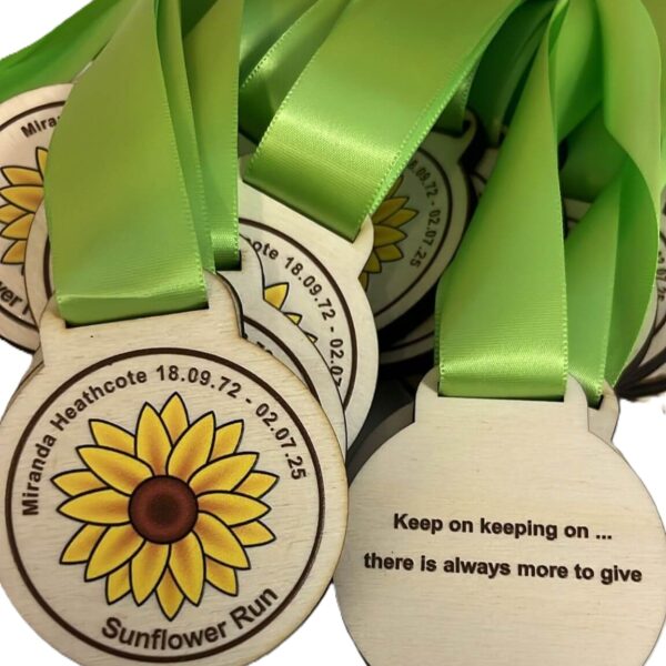 A pile of coloured wooden medals with green ribbons. The medals feature a sunflower, the text “Miranda Heathcote 18.09.72 - 02.07.25 Sunflower Run,” and the phrase “Keep on keeping on ... there is always more to give.”.