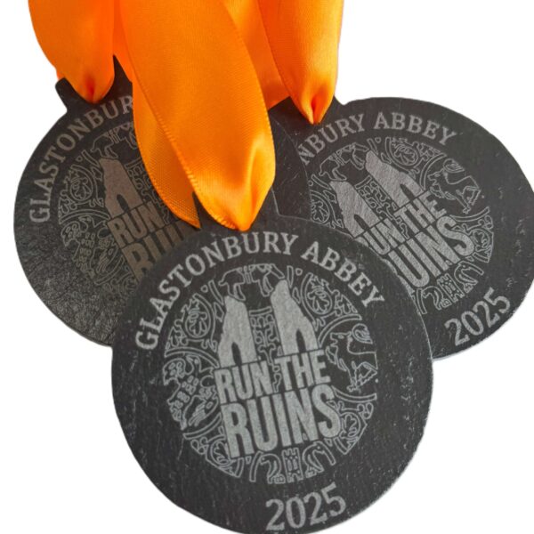 Three black Eco-Friendly Slate Medals with orange ribbons display Glastonbury Abbey 2025 and Run the Ruins, featuring an outline of the abbey ruins and decorative patterns.